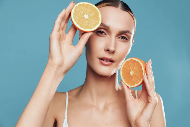 Young woman with clear skin holding two halves of an orange near her face. Blue background and natural light create a fresh and healthy skincare concept.