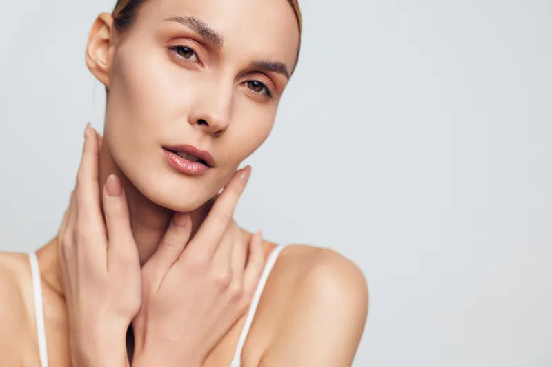 Closeup portrait of young woman with clear skin touching neck gently, neutral expression, soft natural light, minimal makeup, smooth complexion, studio background.