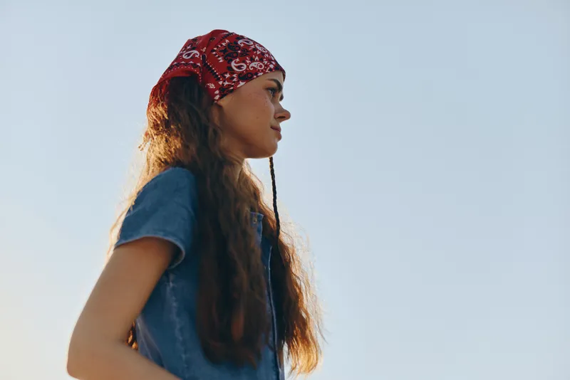 Young woman with long hair wearing red bandana and blue shirt looking to side under clear blue sky on sunny day outdoors.
