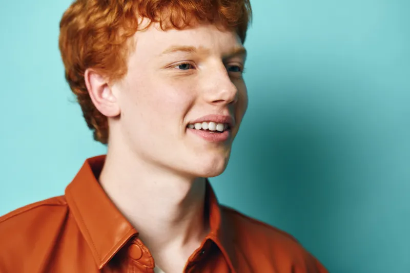 Young man with red hair and fair skin wearing a brown leather jacket smiling in front of blue background. Portrait, casual style and confident mood for lifestyle and fashion.
