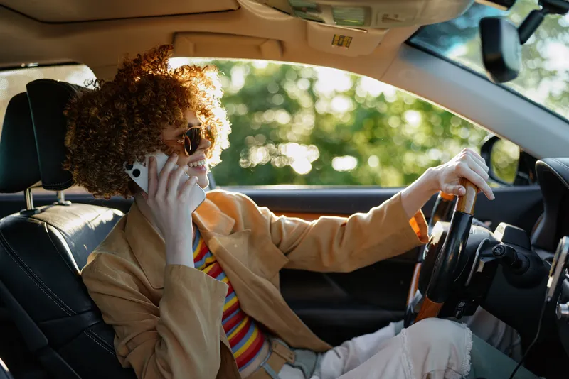A young woman with curly hair drives a car while talking on the phone, wearing a striped shirt and tan jacket, captured with retro film color and warm sunlight.