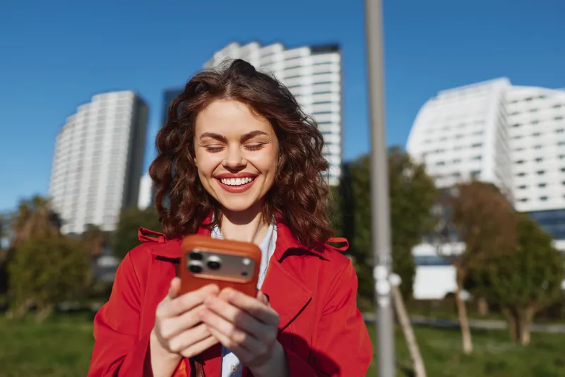 A smiling woman in a red jacket uses a smartphone outdoors with city buildings in the background, capturing a candid moment of everyday life and connection.