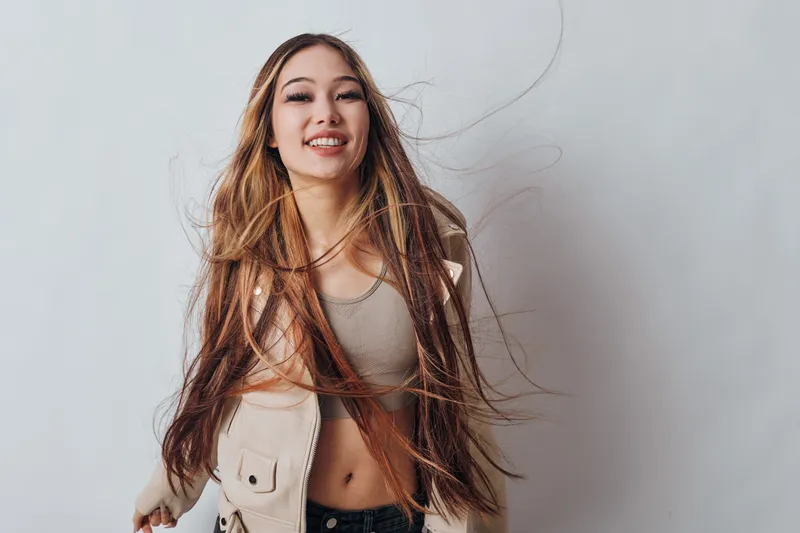 Young woman smiling with long flowing hair jumping in casual clothing against white background. Happy female expressing joy with natural movement and carefree attitude.