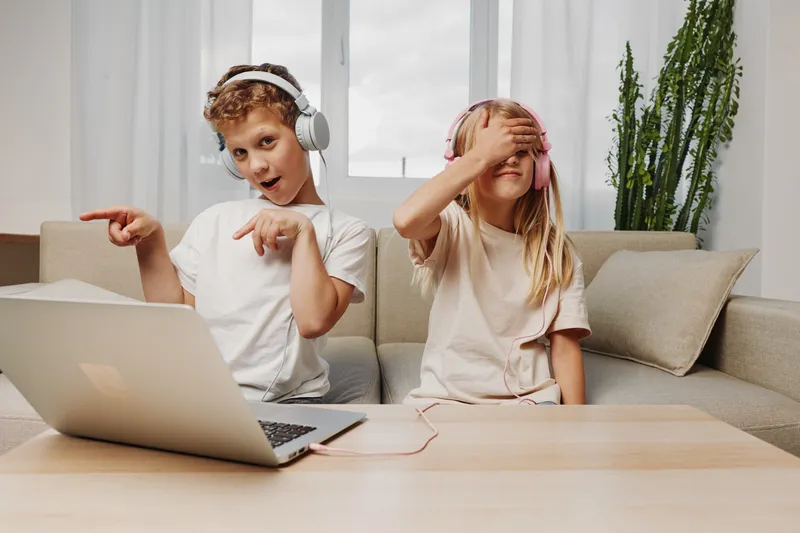 Two children wearing headphones enjoy music and playfully interact with a laptop on a wooden table in a bright living room. Concept of fun, technology, and childhood entertainment.