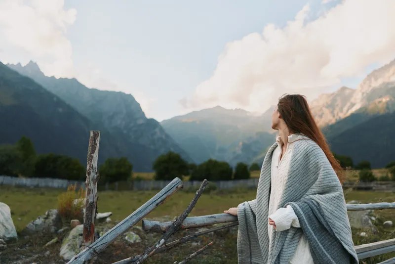 Woman in a cozy shawl stands beside a rustic fence in a valley with distant mountains, conveying a serene lifestyle moment, connection with nature and quiet reflection