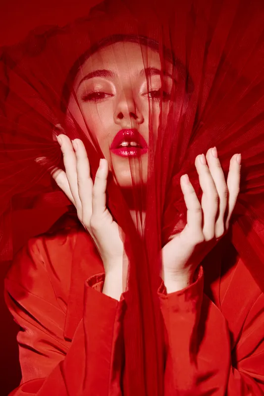 Fashion model wearing vibrant red fabric veil and elegant lipstick poses creatively with hands framed around silk material. Studio beauty portrait in intense red tones. Concept of fashion, beauty,