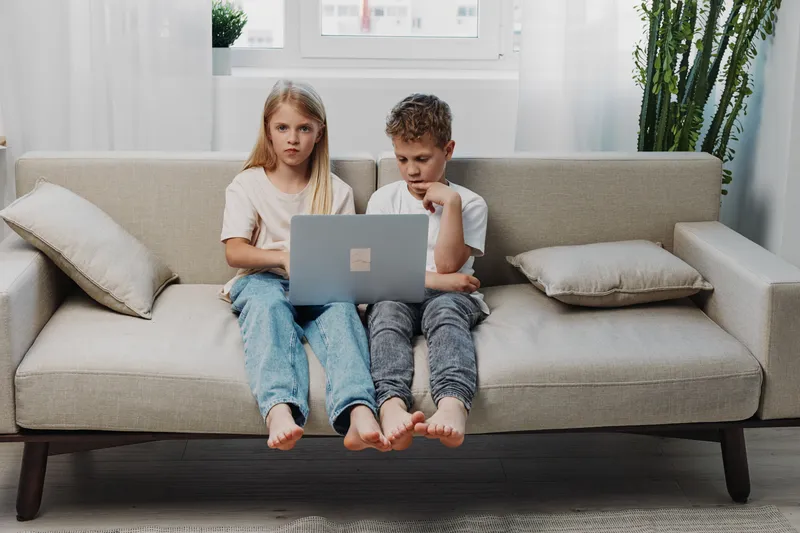 Two children sitting barefoot on a modern sofa using a laptop together in a bright living room. Concept of childhood, technology, learning, and family time at home.