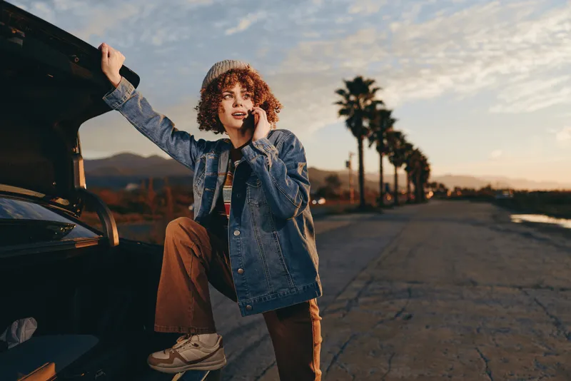 Woman lifestyle scene at sunset, standing by car with open trunk, wearing denim jacket and jeans, curly hair blowing, relaxed confident pose, outdoor road view, warm light