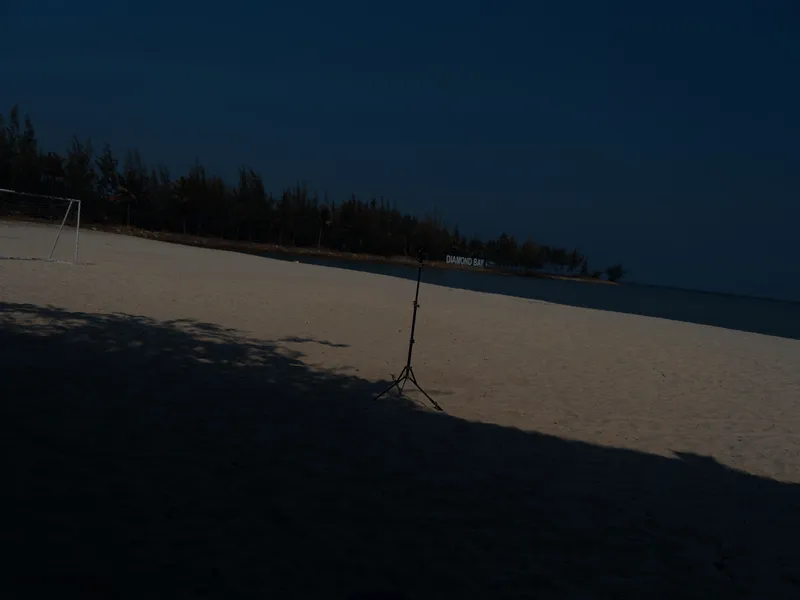 empty sandy beach at dusk with a single light stand casting shadows, calm sea in the background and a goalpost on the left side, peaceful outdoor scene