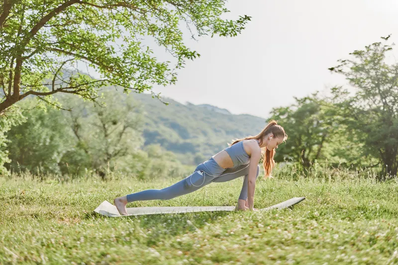 Young woman practicing yoga in a serene outdoor setting, performing a lunge pose on a mat in nature, bright colors and calm emotions, conveying health and wellness