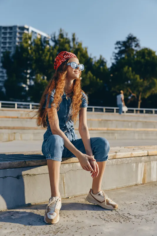 Young woman sitting outdoors wearing casual denim clothes, red bandana, and sunglasses enjoying sunny day with relaxed attitude in urban environment.