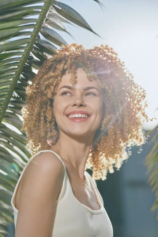 smiling woman with curly hair enjoying sunny day outdoors, green palm leaves background, natural light highlighting radiant skin and joyful expression