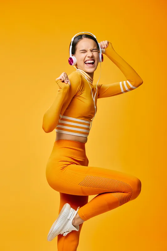 Energetic woman fitness enthusiast jumping midair in bright orange athletic wear with headphones, vibrant studio backdrop, capturing dynamic movement, athleticism and bold workout energy.