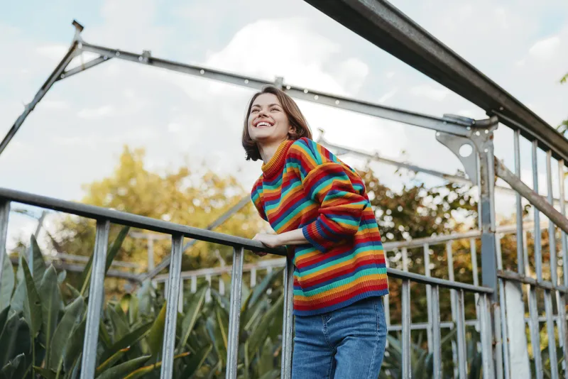Young woman in a colorful striped sweater leaning on metal railing, smiling and looking up in a glass greenhouse, casual denim jeans and short hair, joyful travel lifestyle portrait.