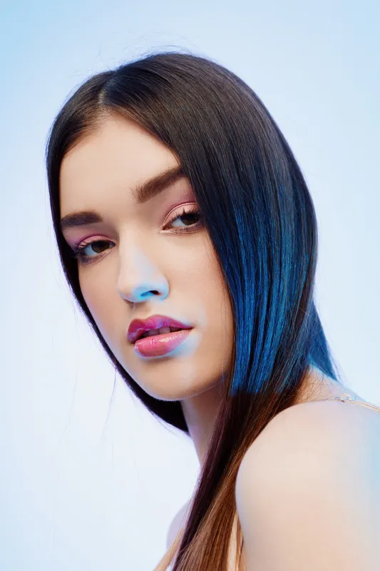 Close-up portrait of a young woman with long dark hair and pink makeup looking at the camera. Studio shot with soft lighting on a light background. Concept of beauty fashion.