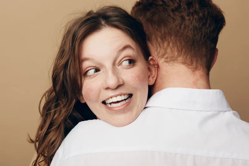 Happy young woman with freckles smiles while hugging a man in white shirt. Intimate moment, love, and affection captured on beige background in casual lifestyle concept.