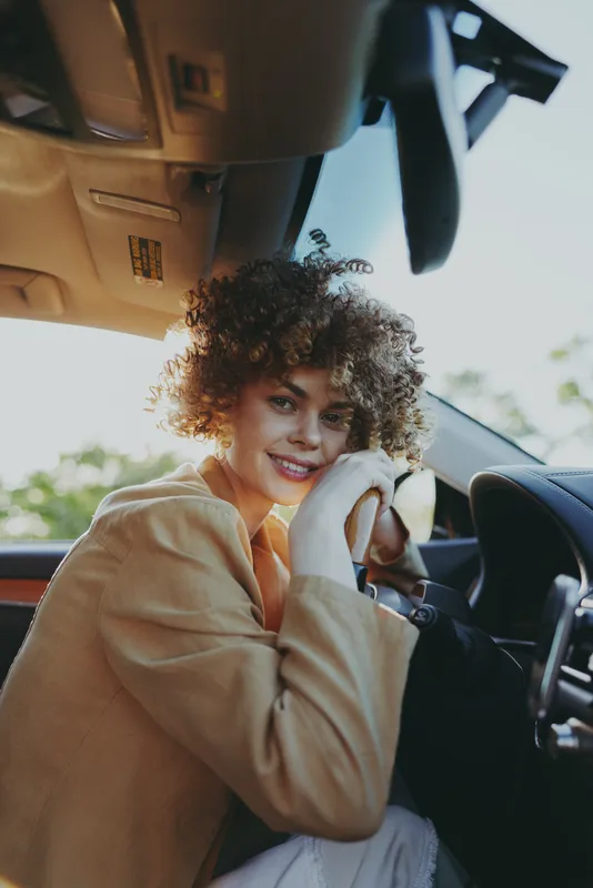 A cheerful woman sits in the driver's seat of a car, wearing a beige jacket, capturing a relaxed lifestyle moment in warm retro film color tones.