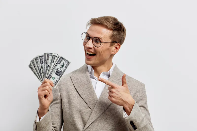 excited man holding fan of dollar bills pointing finger smiling in beige jacket smart casual finance concept cryptocurrency digital wealth