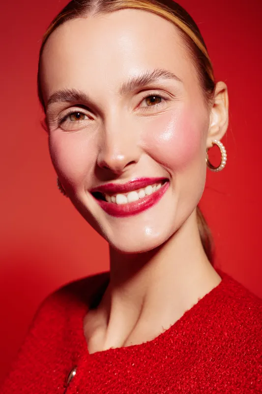 Smiling woman with clear glowing skin and red lipstick poses against red background wearing red sweater and hoop earrings. Concept of beauty, skincare, fashion, and confident female portrait.