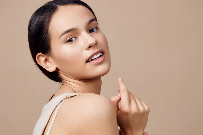 Young woman with a serene expression looking back over her shoulder against a neutral background, showcasing natural beauty and smooth skin