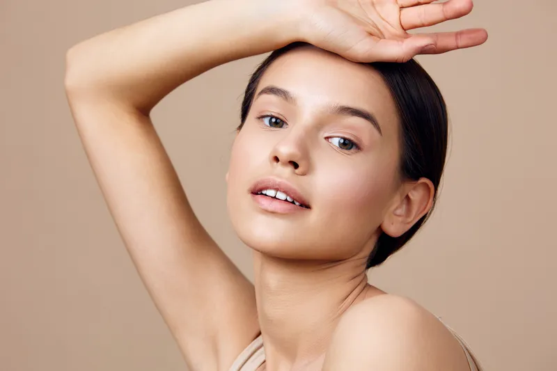 Beautiful young woman with natural makeup posing gracefully against a neutral background, showcasing her radiant skin and confident expression