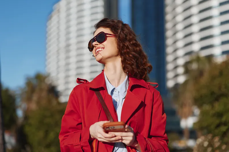 A stylish woman in a red coat smiles outdoors in an urban setting, wearing sunglasses and holding a phone. Concept: casual confidence in city life.
