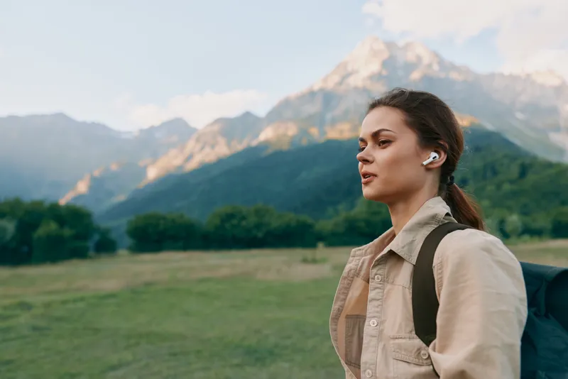 Young woman hiking outdoors with a backpack and wireless earbuds, gazing ahead as rugged mountains rise behind her, capturing a calm travel moment