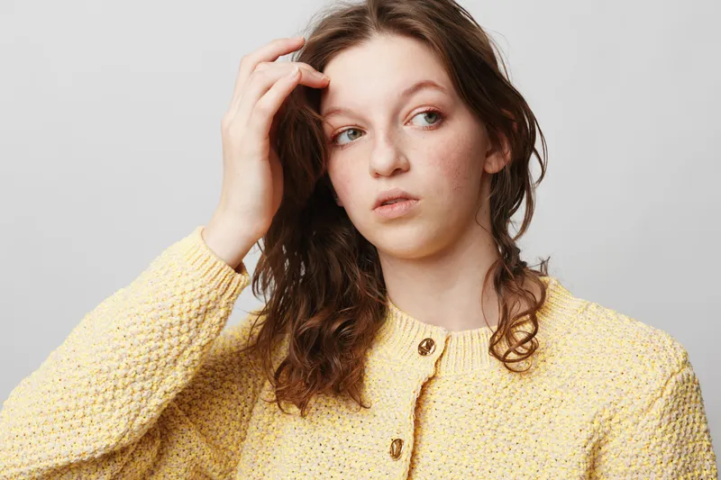 Young woman with curly hair wearing yellow knitted sweater looks away thoughtfully. Studio portrait on light background. Concept of contemplation, casual fashion, and natural beauty.