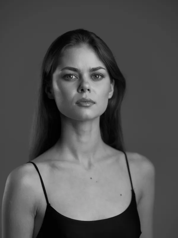 Black and white portrait of serious woman with natural makeup and dark hair wearing thin strap top, minimalistic studio background
