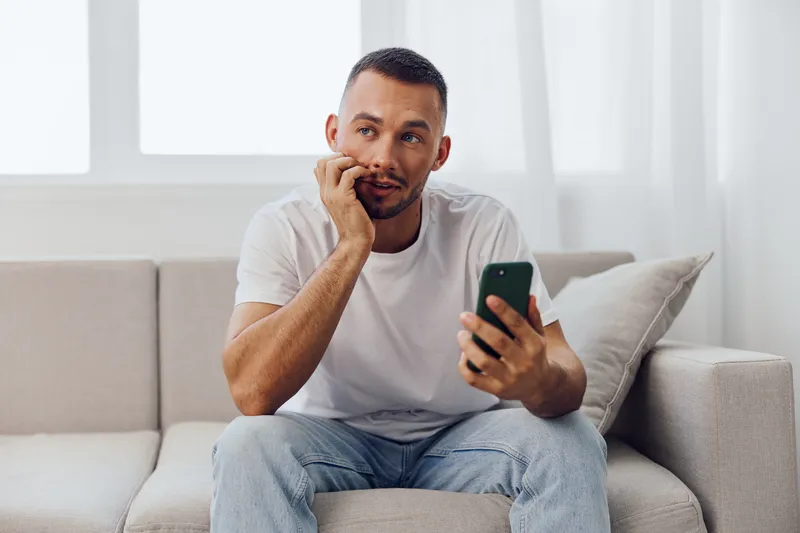 Thoughtful man sitting on a couch, holding a smartphone, wearing a casual outfit, lost in contemplation, natural light streaming through the window.
