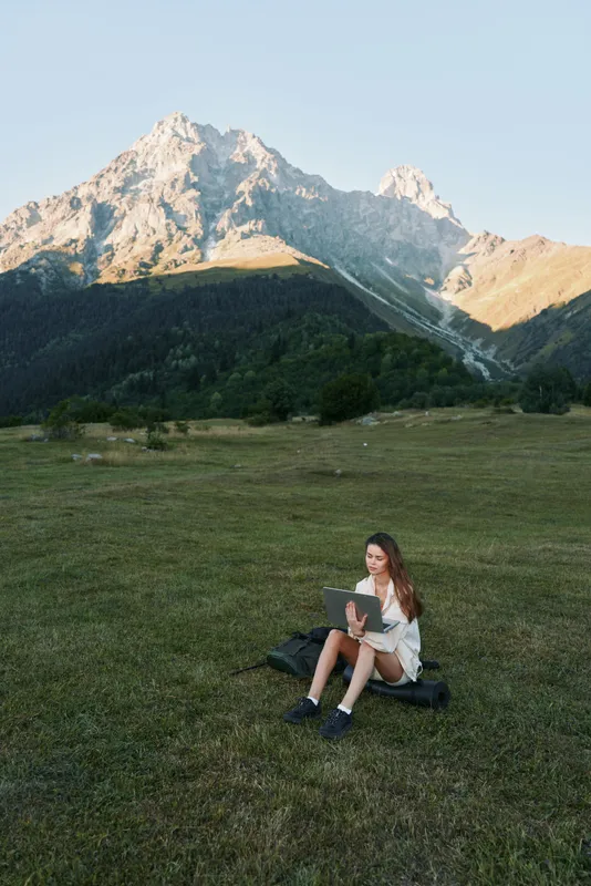 A young woman sits on a grassy meadow with a laptop in hand, surrounded by towering mountains. She works outdoors, appearing focused and calm in a tranquil natural setting.