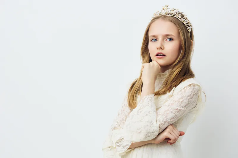 Young girl in elegant white dress with crown, looking confidently at the camera, conveying a sense of royal beauty and innocence against a minimalist background