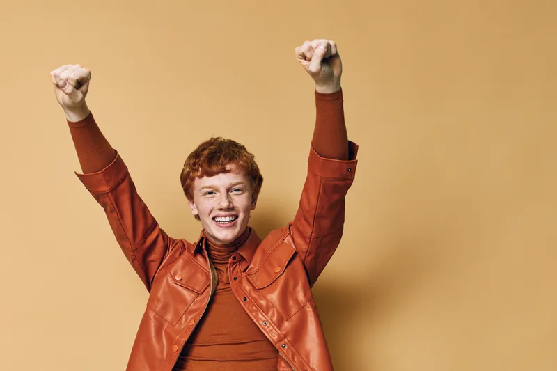 Young man with red hair wearing orange jacket raising arms in excitement against beige background. Smile, celebration, happiness and casual style with positive emotion and victory gesture.