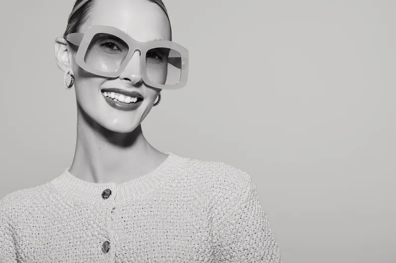 Smiling Caucasian woman with light skin wearing large stylish sunglasses and a textured sweater. Portrait with confident expression and clean background for fashion and beauty concept.