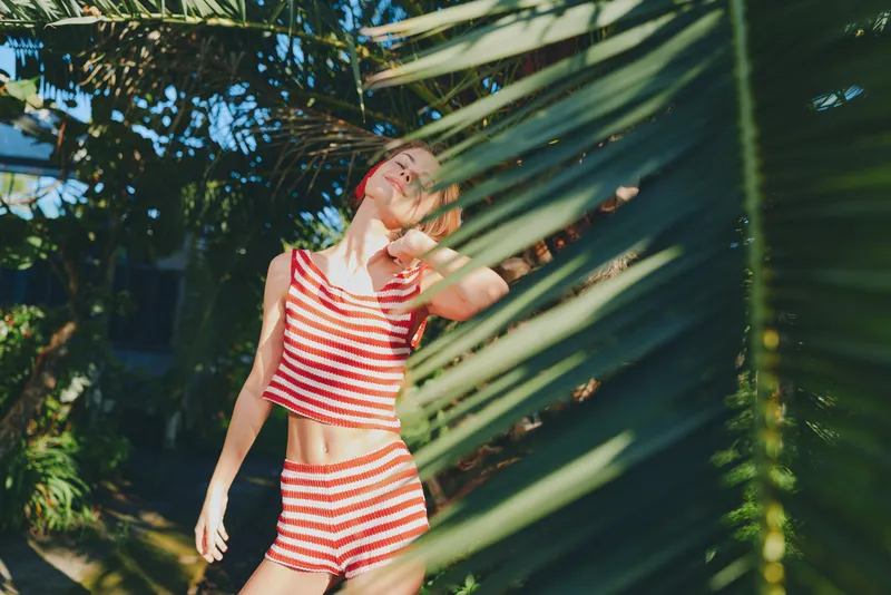 A cheerful person in bright striped swimwear stands amid lush tropical plants, enjoying sunny weather and carefree moment, creating a vibrant summer scene.