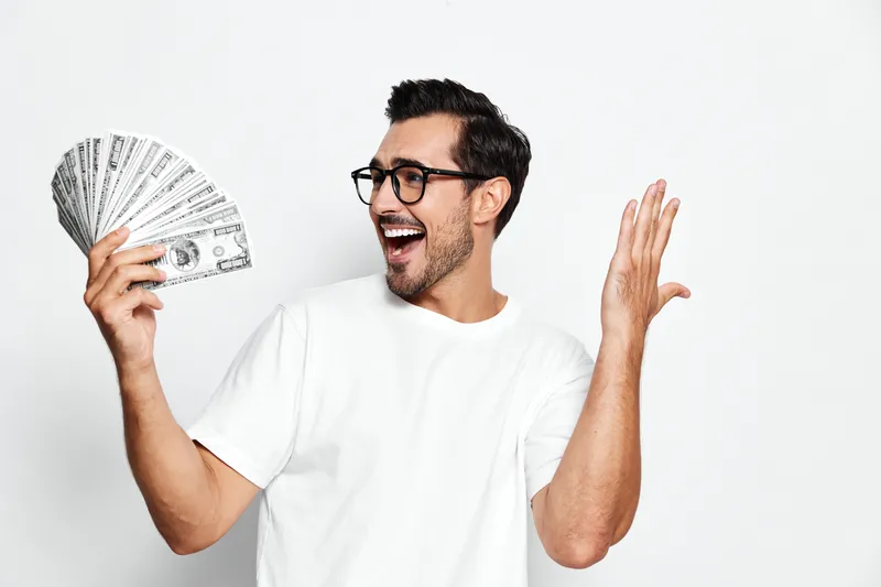 Happy smiling man in white t-shirt holding cash money and celebrating his success with joy. Isolated on plain background showcasing excitement and achievement.
