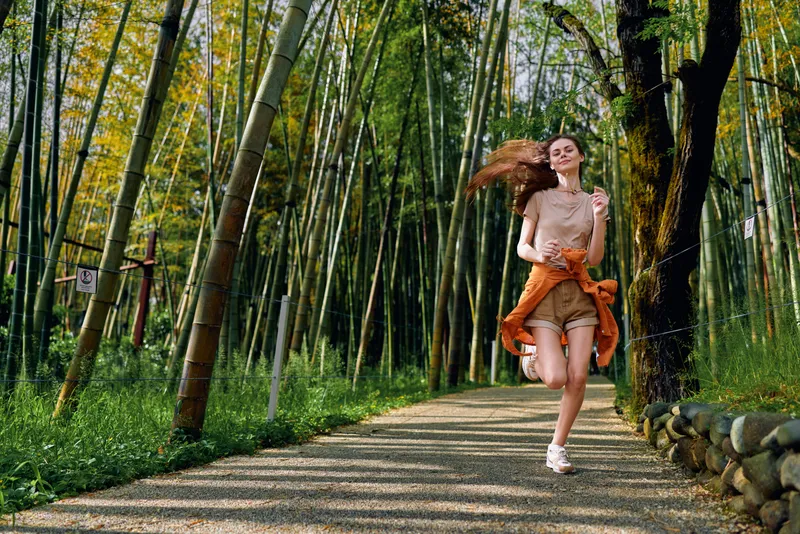 Woman running along a bamboo path in a serene forest, smiling and enjoying outdoor jogging in a casual summer outfit with sweater tied at waist, motion and natural light.