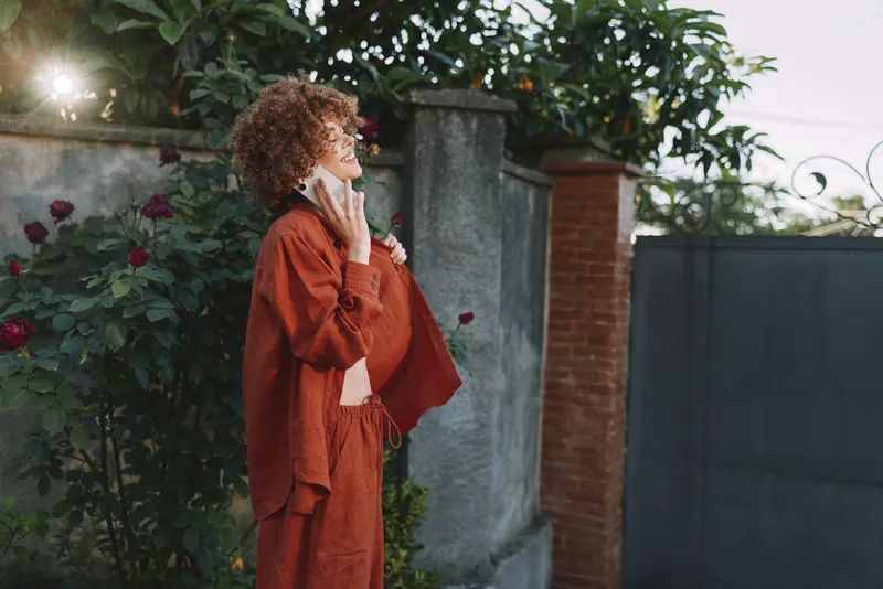 Vibrant woman outdoors, wearing red outfit, thoughtful pose beside a wall, warm sunlight highlights textured fabrics, lush greenery backdrop, contemporary lifestyle moment