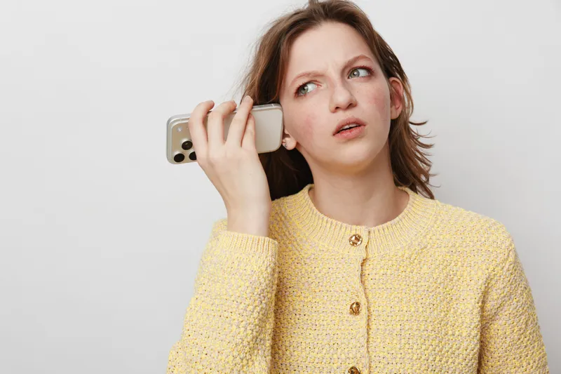 Confused young woman in yellow sweater holds smartphone to ear, looking puzzled. Isolated on white background. Concept for misunderstanding, communication problems, and technology frustration.