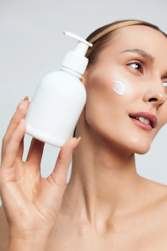 A young woman with clear skin holds a white lotion bottle near her face with a cream dot on her cheek. Concept for skincare, beauty routine, and moisturizing products.