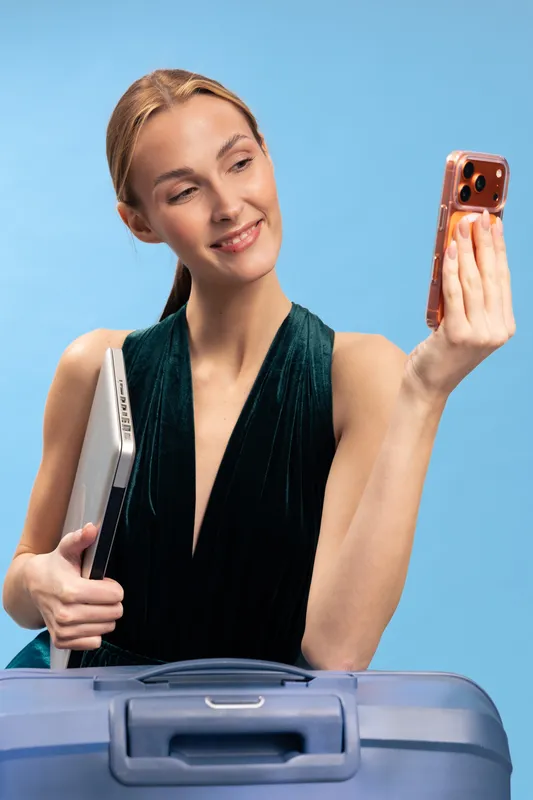 Young woman with laptop and suitcase takes selfie with smartphone on blue background. Business travel concept, smiling female traveler ready for trip, modern technology and vacation mood.