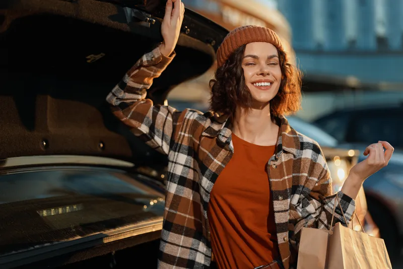 Woman shopping by car trunk with a warm smile in urban golden hour light, candid lifestyle portrait conveying authenticity, mindful living and emotional storytelling in a cozy city moment.