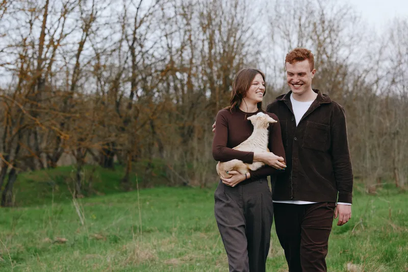 Young couple walks through a grassy field holding a baby goat while smiling and enjoying a peaceful moment together in casual autumn clothing.