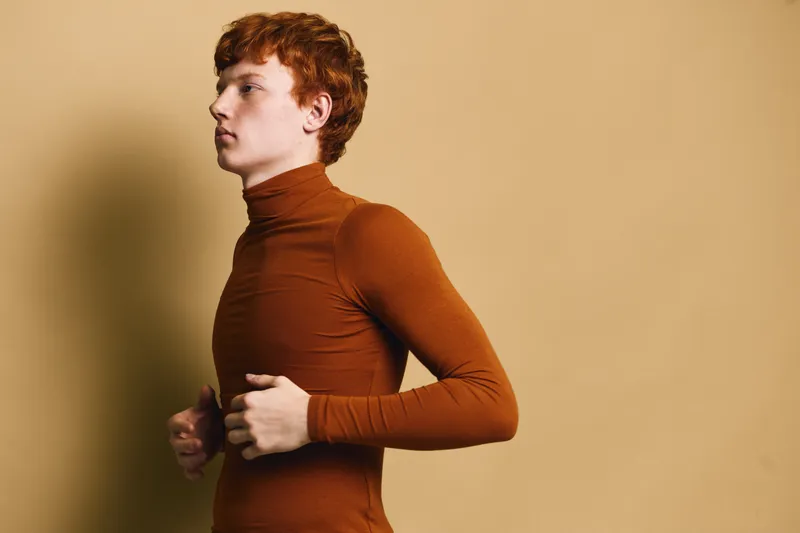 Young man with red hair in a brown turtleneck sweater posing sideways on beige background. Studio portrait of male model with serious expression and casual outfit for fashion.