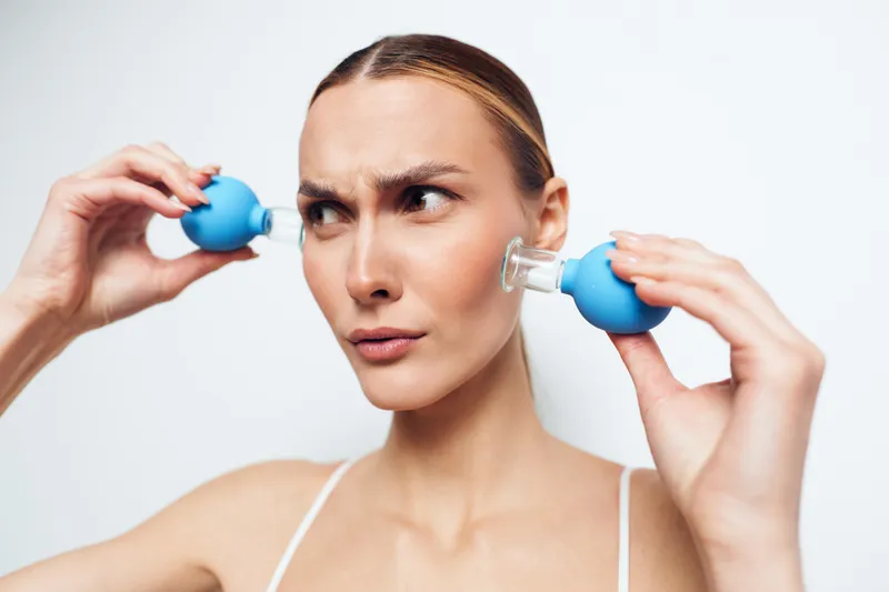 Young woman with serious expression using blue facial suction cups on cheeks. Skincare concept with close-up on white background and soft natural lighting for beauty routine.