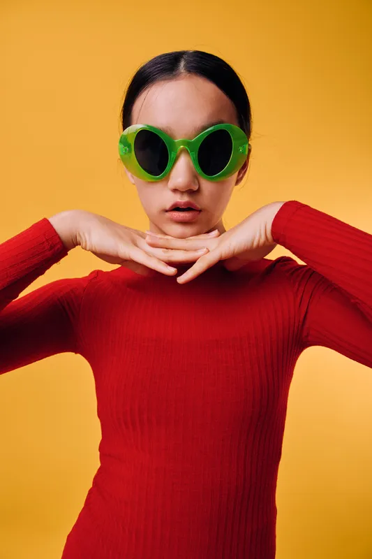 Young woman wearing large green sunglasses and red sweater posing with hands under chin against yellow background. Fashion, style, portrait, casual outfit, confident expression with modern