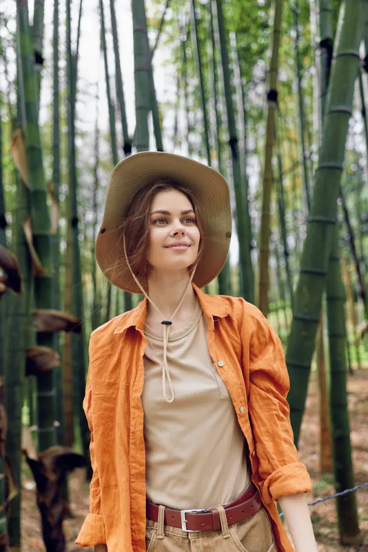 Woman in hat stands among tall bamboo in a forest, portrait with a gentle smile, outdoor nature lifestyle scene, casual orange shirt and straw hat conveying travel and serene adventure.