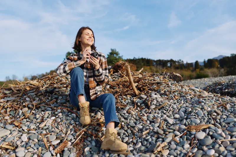Woman in plaid shirt and jeans sits on a rocky beach, holding a camera, enjoying the outdoors. Bright sky and rugged terrain create a relaxed lifestyle portrait with nature and travel vibes.