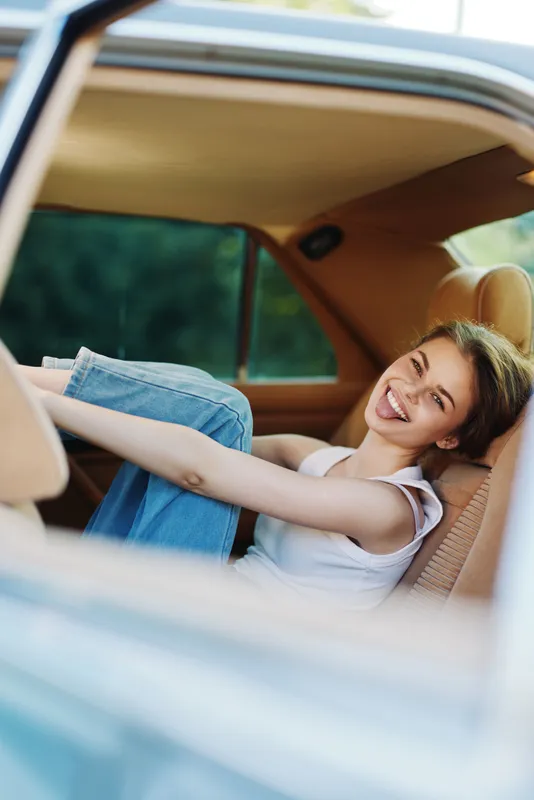 Young woman smiling happily in a vintage car, wearing casual outfit with jeans, embodying joy and freedom, against a vibrant green background.