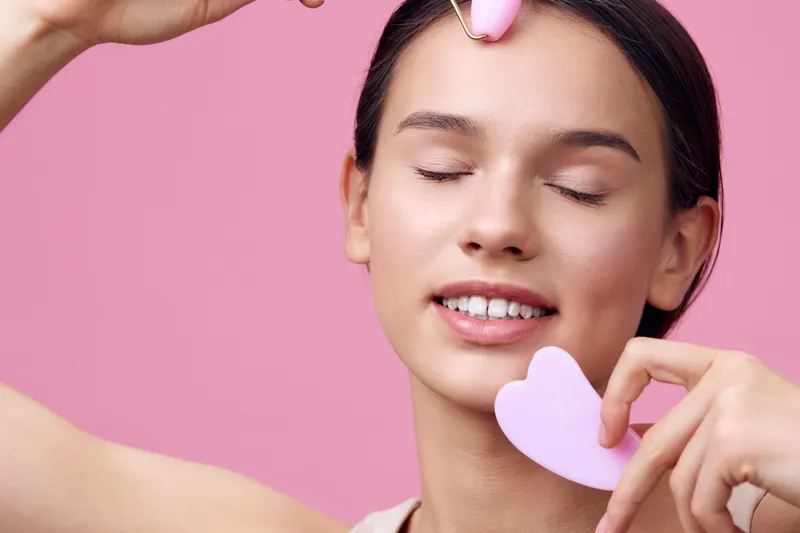 Young woman performing facial massage with pink tools, showcasing relaxation and beauty care in a vibrant pink setting